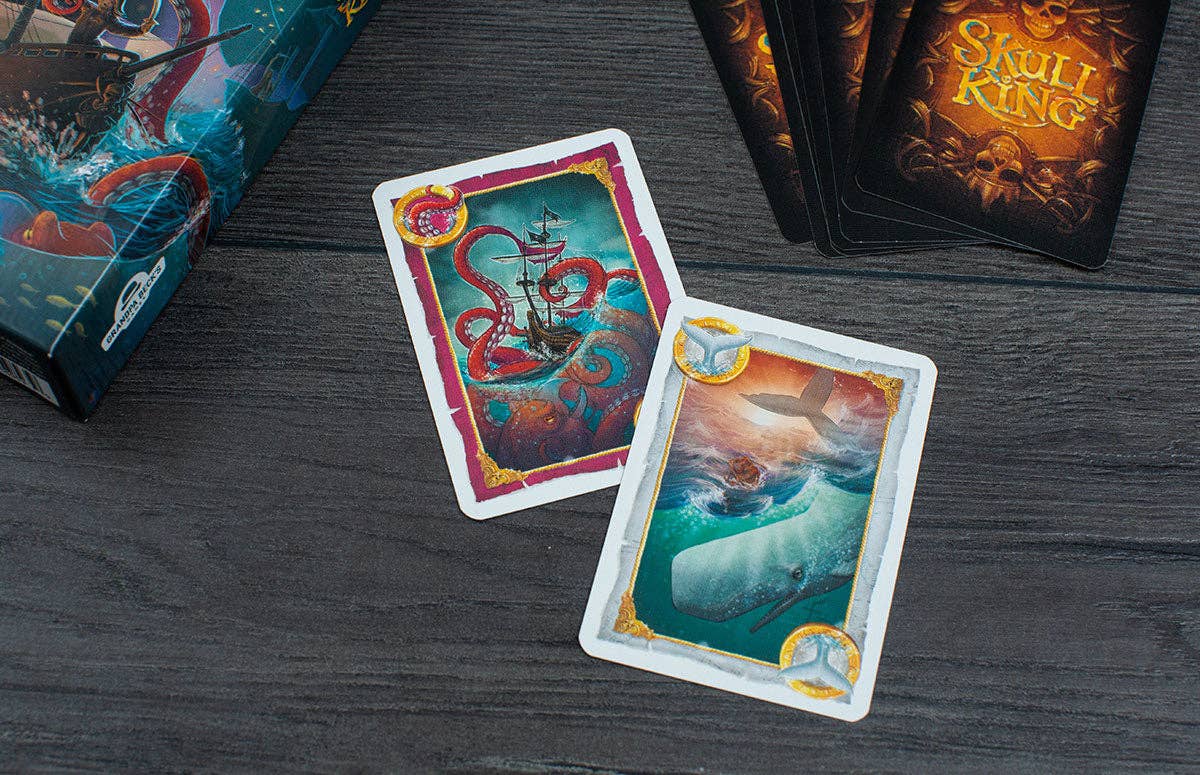 Two colorful playing cards on a dark wooden surface with game boxes in the background.