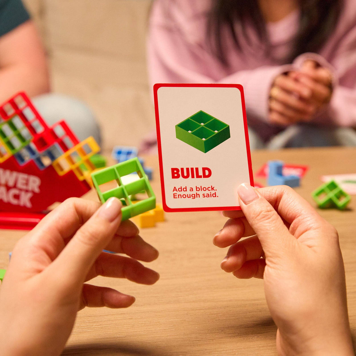 Two hands holding a card with building block instructions and a green block on a wooden table.