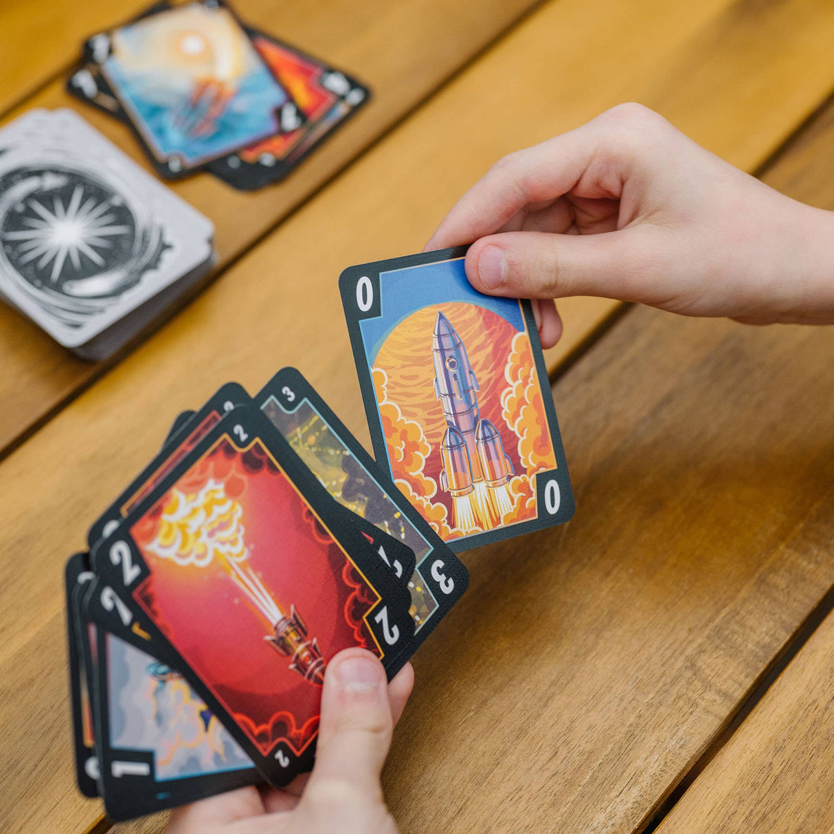 Hand holding a card with a rocket design on a wooden table with other cards.