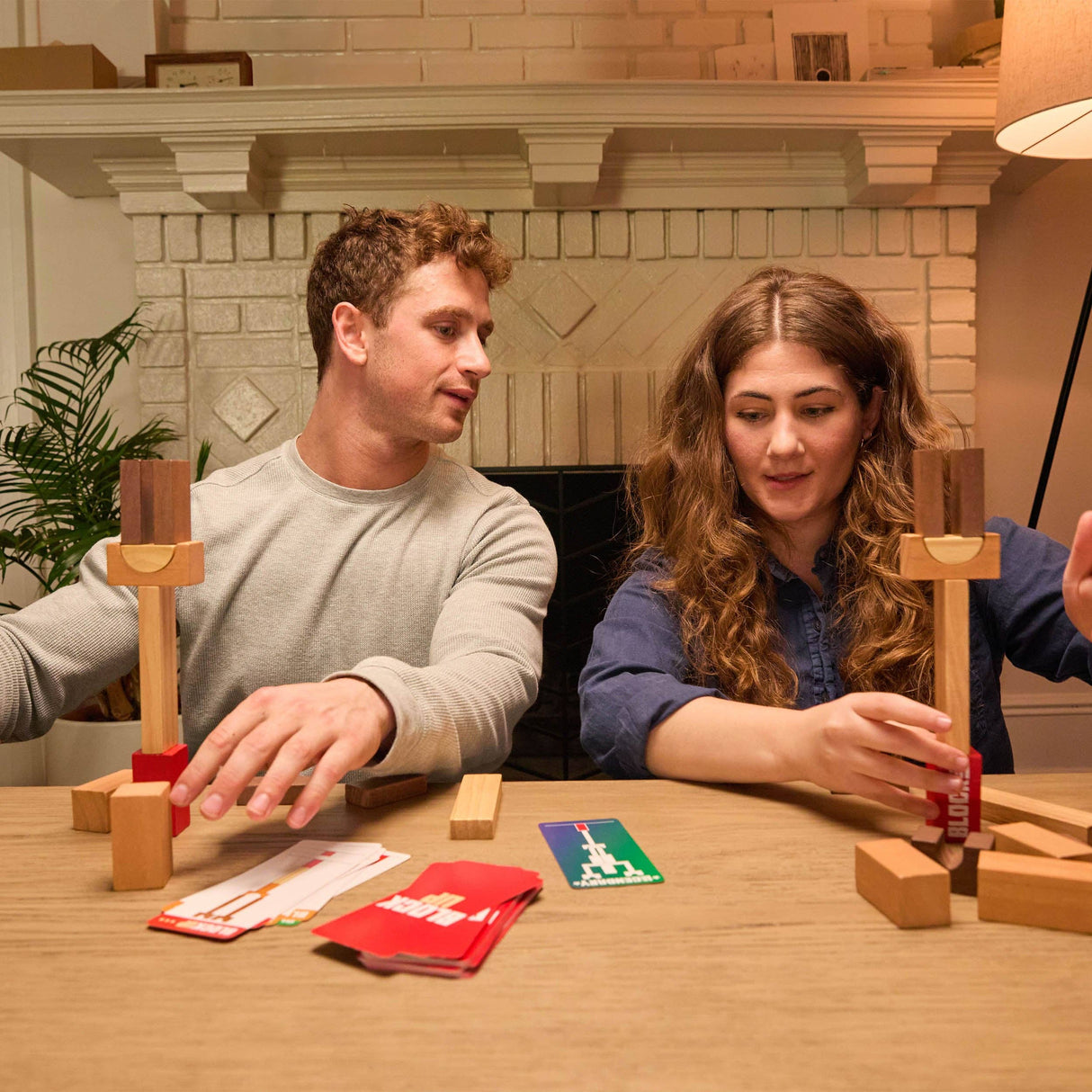 Two people playing a wooden block game at a table with a fireplace in the background.