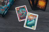 Two colorful playing cards on a dark wooden surface with game boxes in the background.
