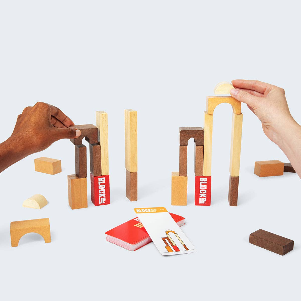 Wooden building blocks with red labels being played with by two hands on a light gray background.
