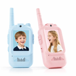 Two toy walkie-talkie style children's phones in pastel blue and pastel pink, shown side by side on a white background. Each device has an antenna, side buttons, a speaker grille, and a screen displaying a child's portrait.