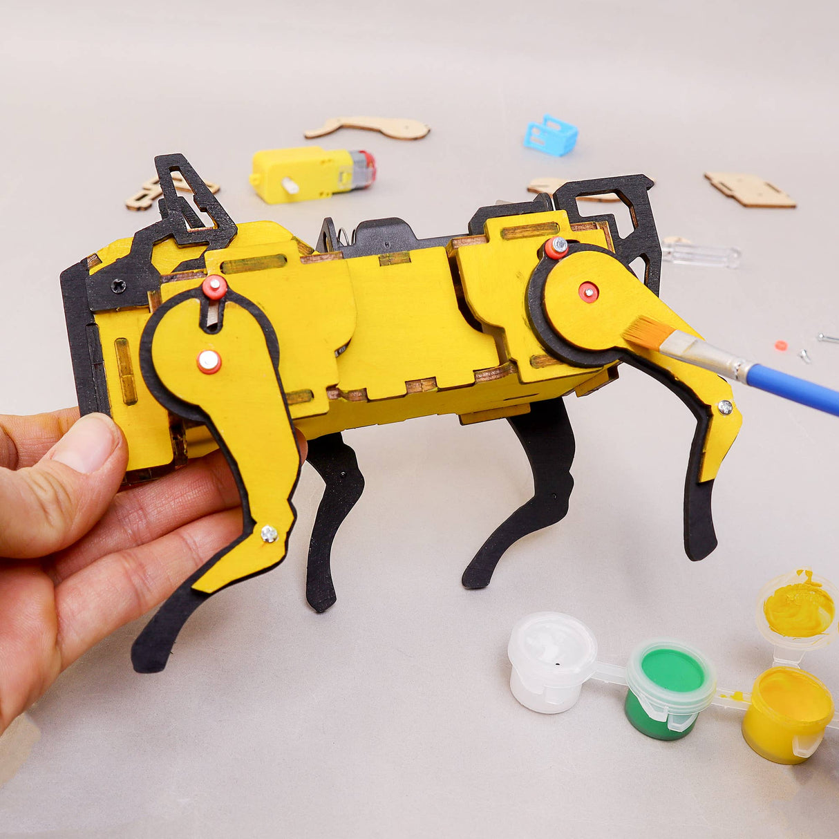 Yellow and black mechanical model kit held by a hand with paint cans and tools on a light gray background.