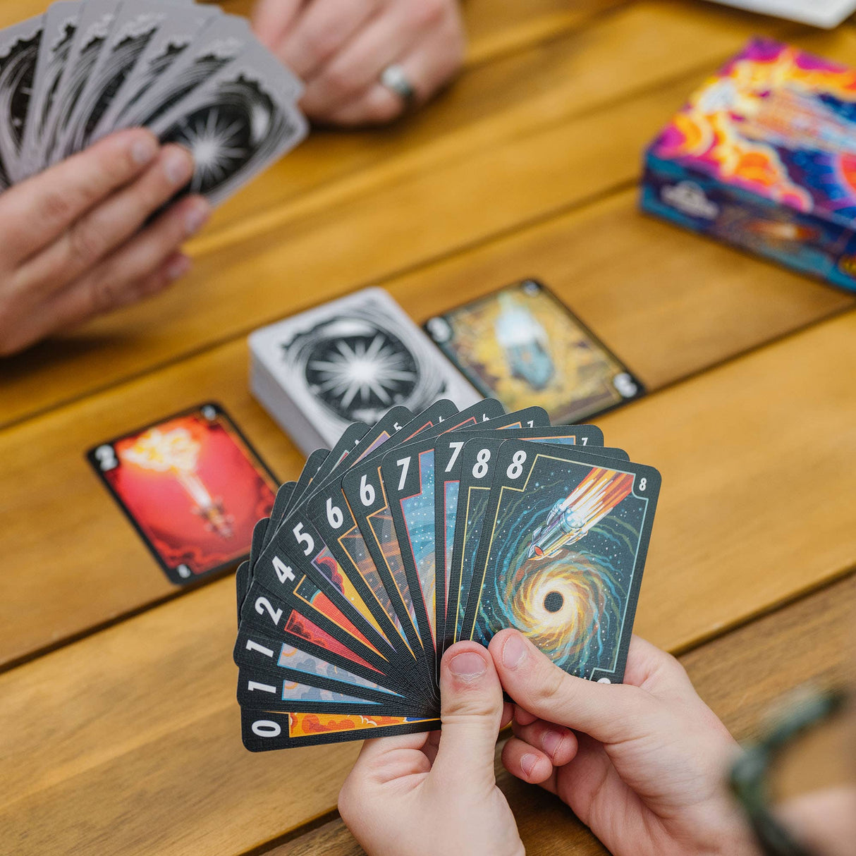 Person holding tarot cards on a wooden table with other cards and a colorful deck in the background.